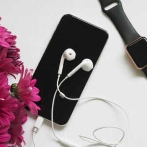 A flat lay of smartphone, smartwatch, and earphones beside vibrant pink flowers on a white background.